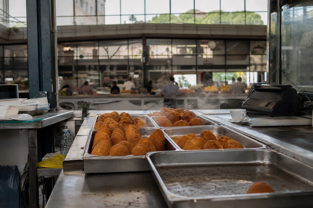 Steam rising from a hot food counter with trays of fried snacks in a Roman market hall