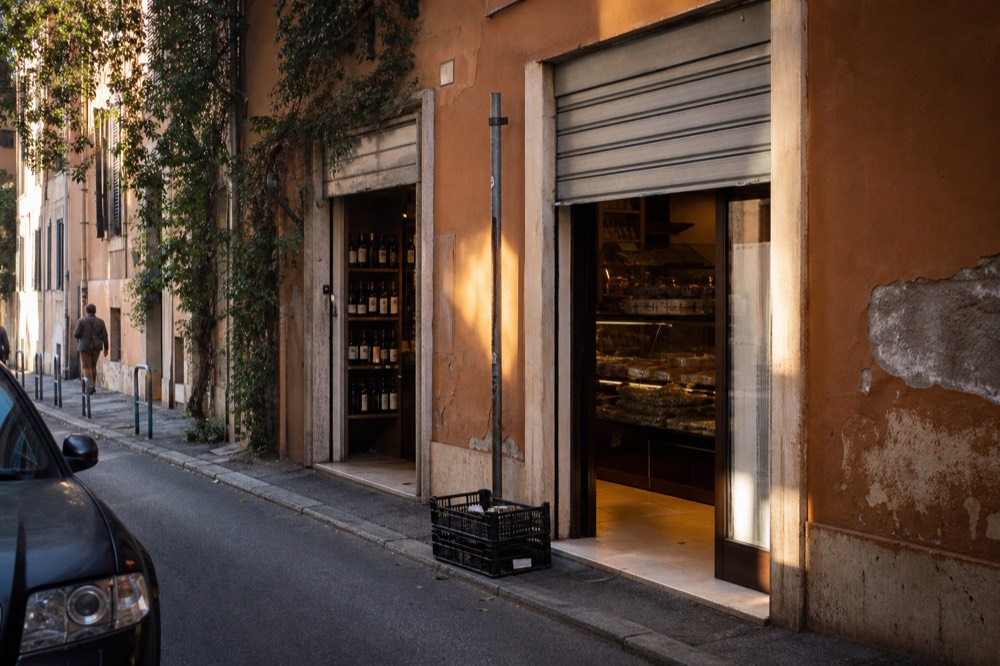 A quiet Roman side street with bakery and wine shop fronts in late afternoon light