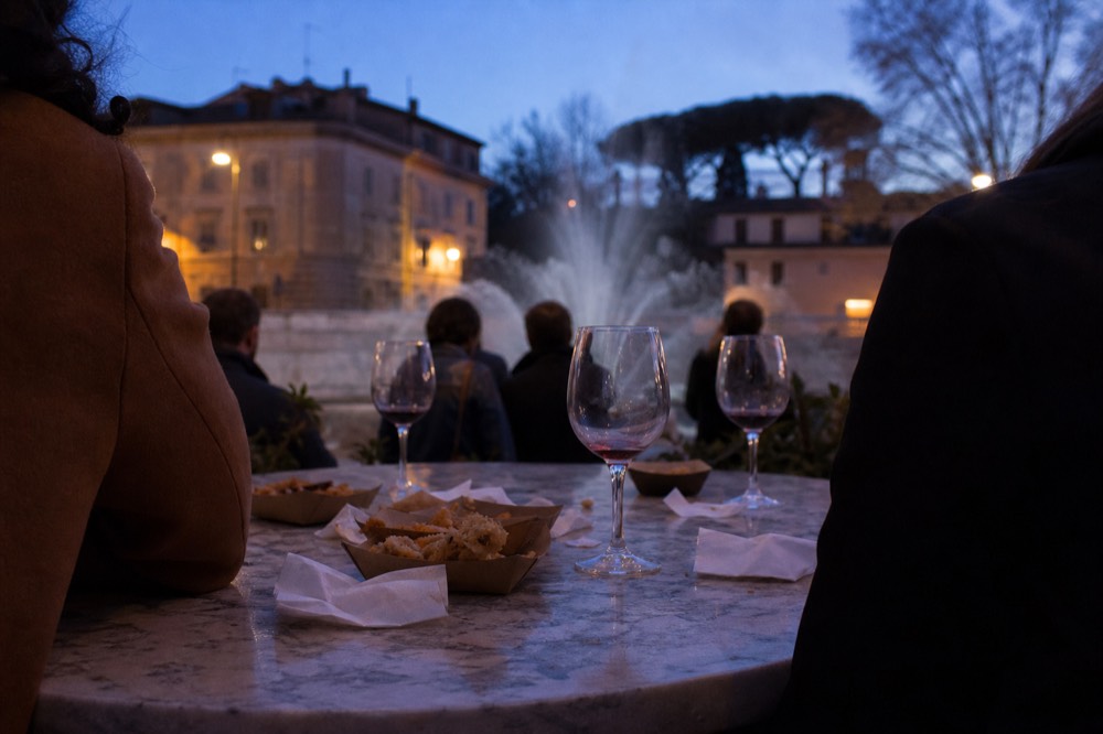 A small group tasting fried snacks and glasses of wine at a high counter in a Roman street at dusk