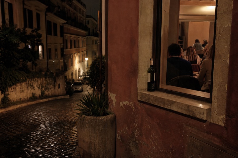 A narrow Roman street at night with warm restaurant light spilling onto cobblestones