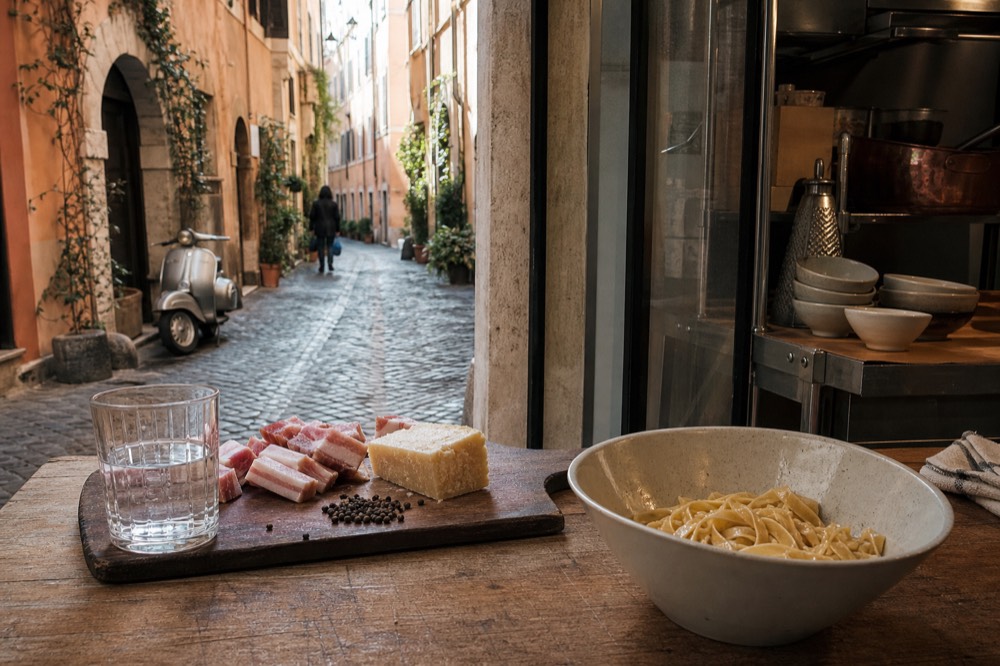 A wooden board with guanciale, pecorino romano and black pepper beside a bowl of pasta