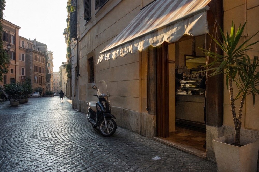 A sunlit Roman street in the morning with a bar awning and counter visible through an open door