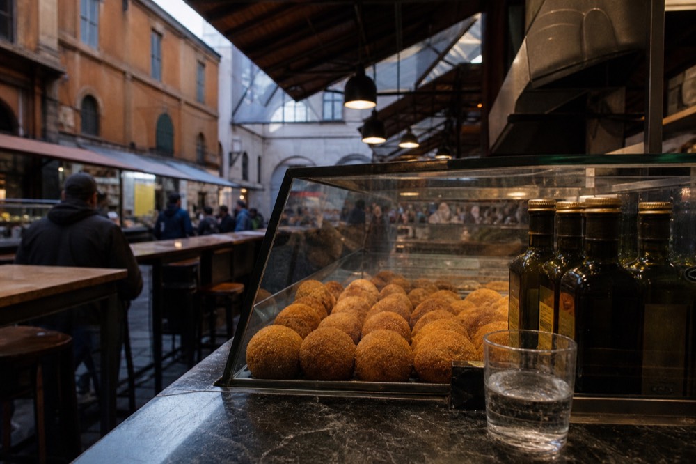 Rows of golden round arancini in a deli display case beside olive oil bottles