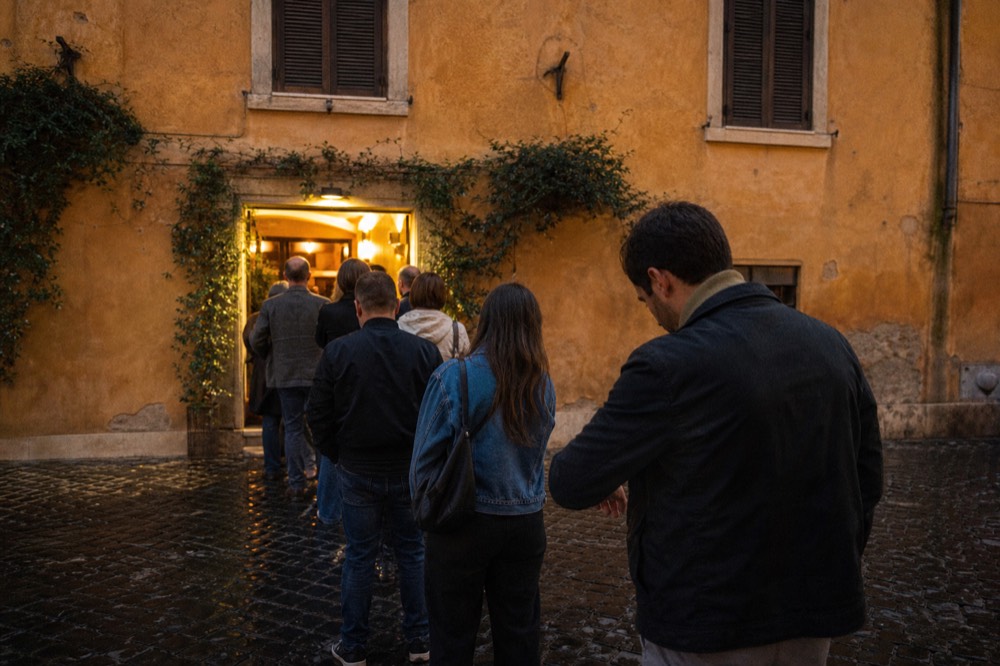 A queue of diners waiting outside a lit trattoria doorway in the evening
