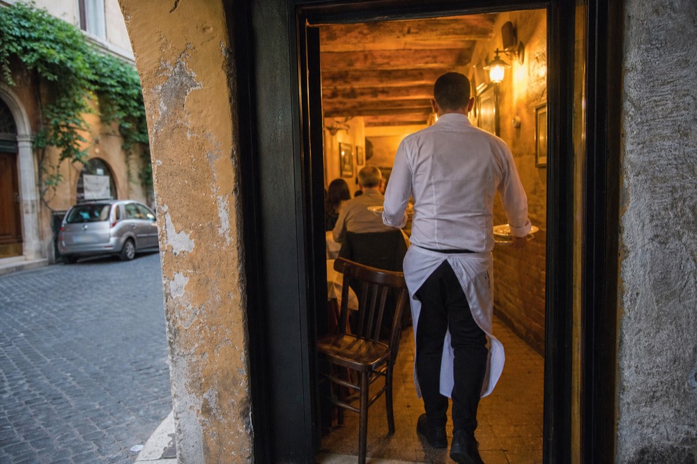 A waiter carrying plates through a narrow Roman trattoria aisle at dinner