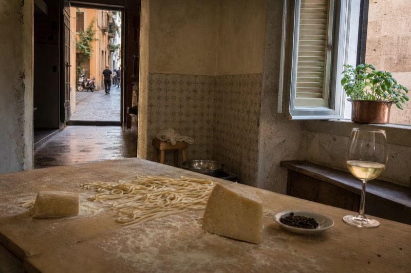 A cook's floury hands shaping tagliatelle on a worn wooden board, a rind of pecorino, a pepper grinder and an open glass of white nearby