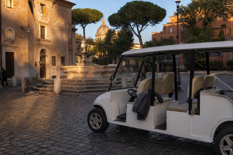 An open electric cart parked on a cobbled Roman square at golden hour, umbrella pines and a weathered fountain rising behind it