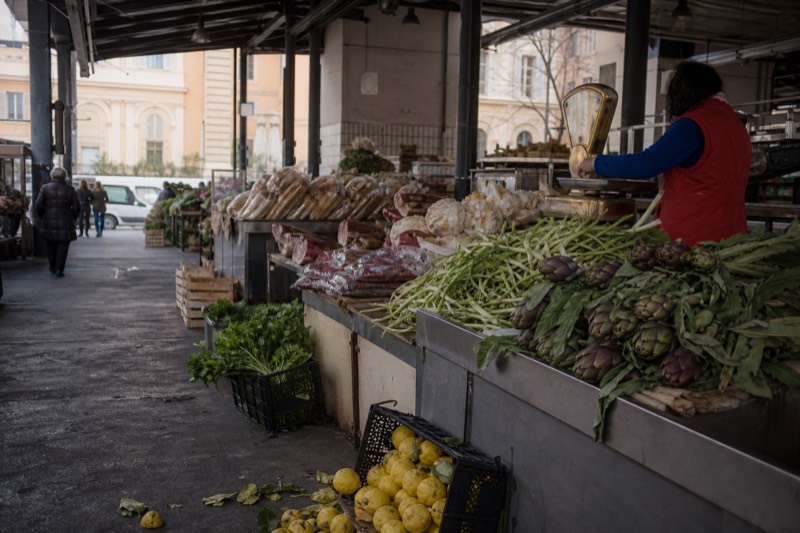 Crates of romanesco, artichokes and salumi beneath the covered stalls of a Rome food market, a vendor handing a wrapped order across the counter