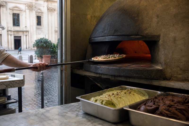 A pizzaiolo sliding a thin Roman base into a deck oven while tubs of stracciatella and pistachio gelato wait on the marble counter