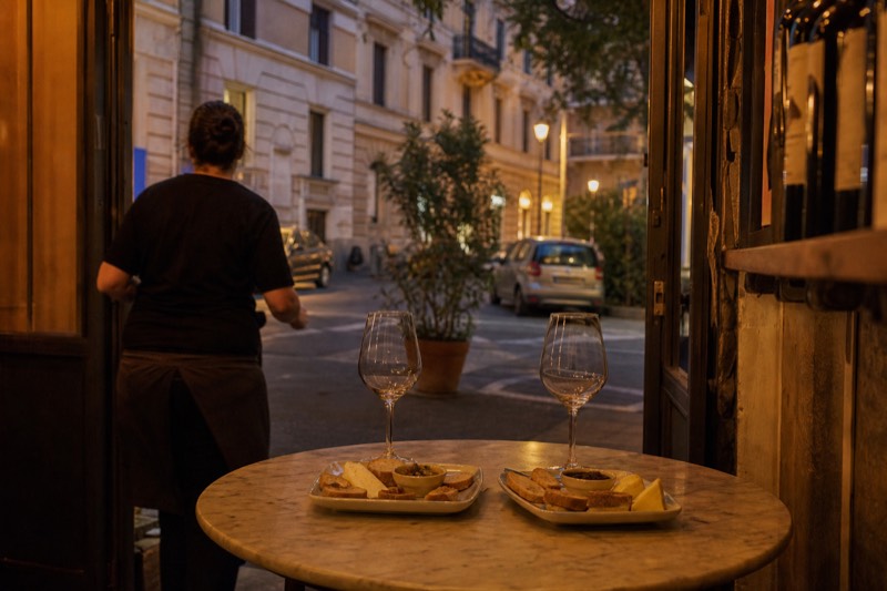 A host arranging bruschetta and two filled wine glasses on a small marble table inside a quiet Roman enoteca, the lamplit street beyond the doorway