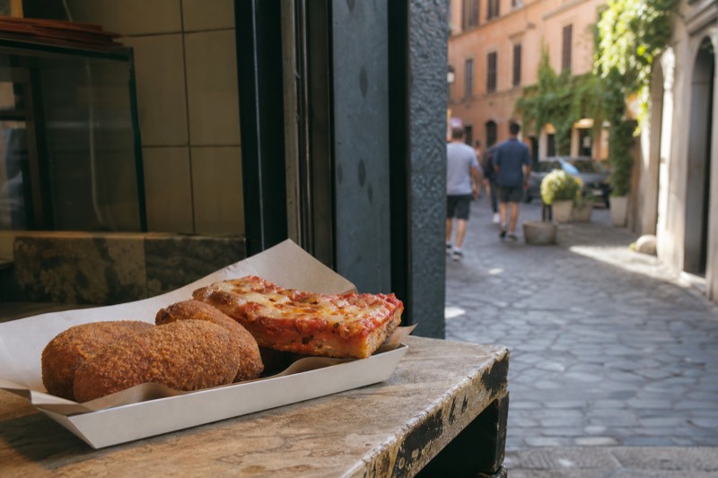 Hands holding a paper cone of hot suppli beside a slice of pizza rossa on a marble counter, the blurred evening street of central Rome visible through the open door