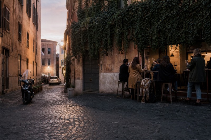 Travellers raising glasses of red wine at a standing counter beneath string lights on a narrow Trastevere street in Rome at dusk