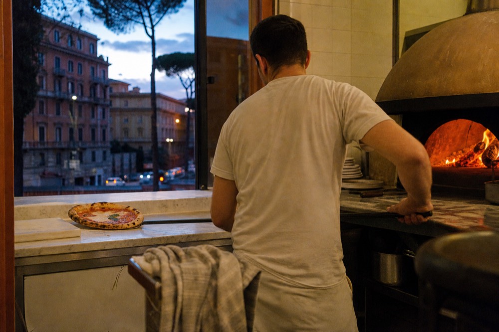 A wood-fired oven glowing behind a pizzaiolo sliding a round pizza onto a peel