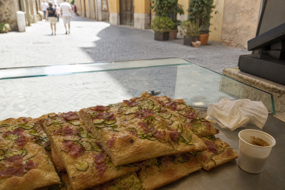 A glass counter of rectangular pizza al taglio slabs topped with vegetables and cured meat