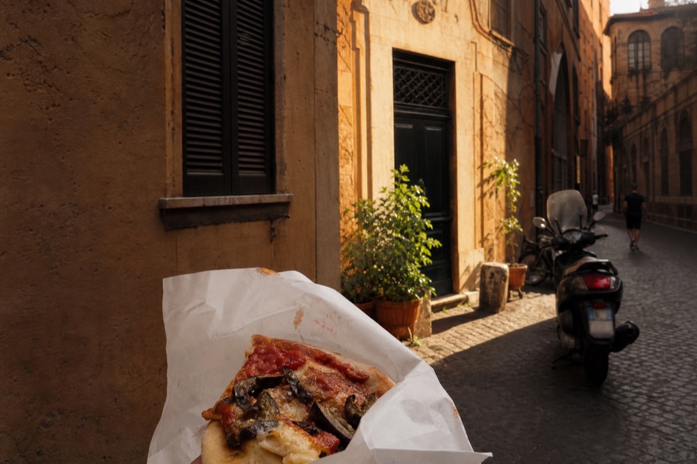 A paper-wrapped slice of pizza al taglio being eaten standing on a Roman street