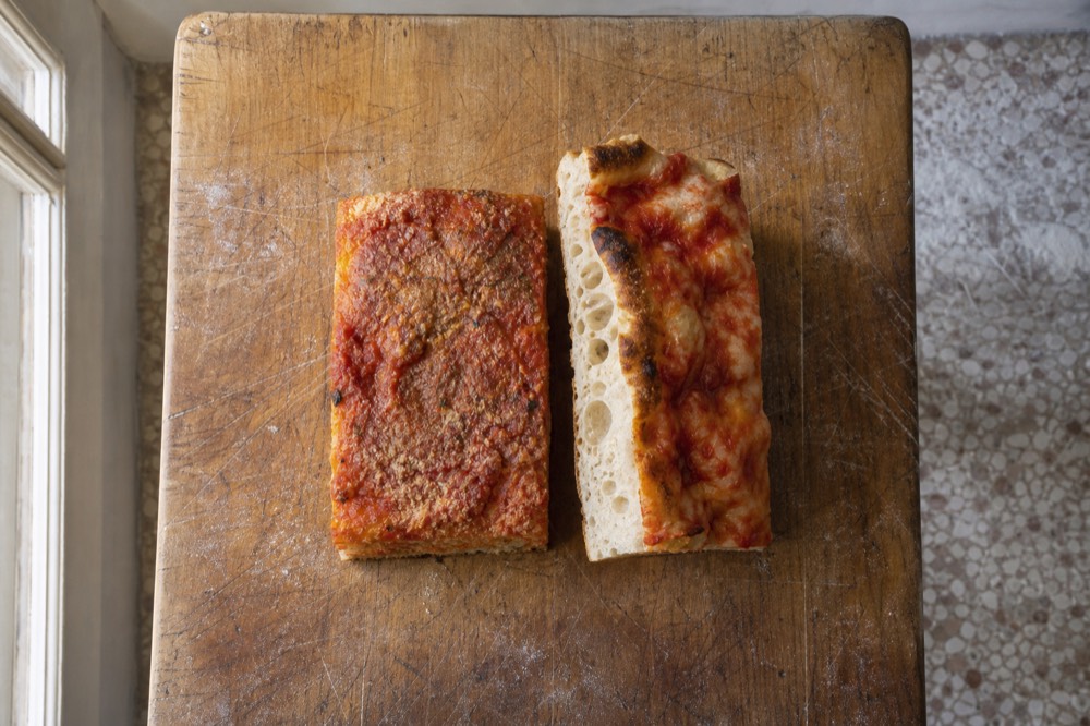 A slab of thick Sicilian sfincione beside a thin Roman al taglio slice on a wooden board