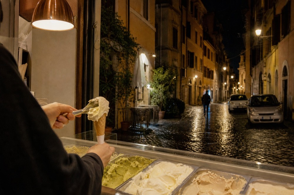 A gelato counter with flat metal tins of muted-coloured flavours and a cone being filled