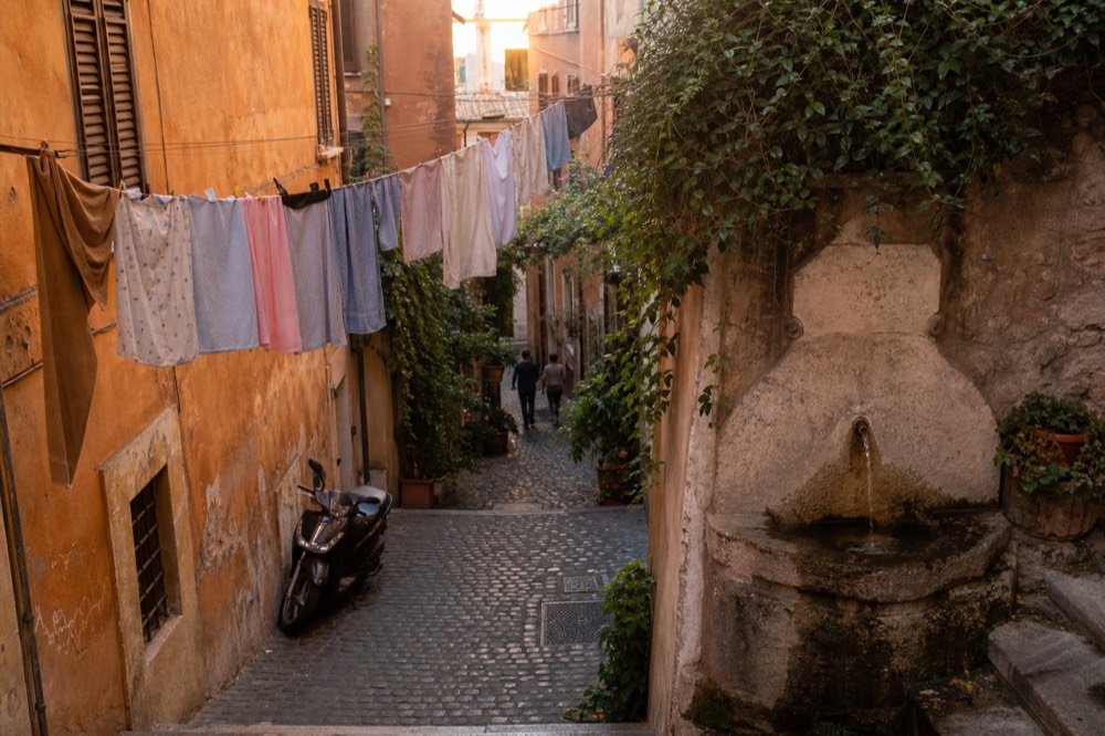 A narrow Roman street with a fountain at the corner and laundry overhead