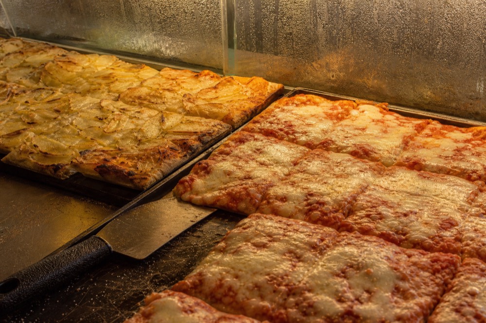 A bakery counter with flat trays of pizza al taglio under the lights