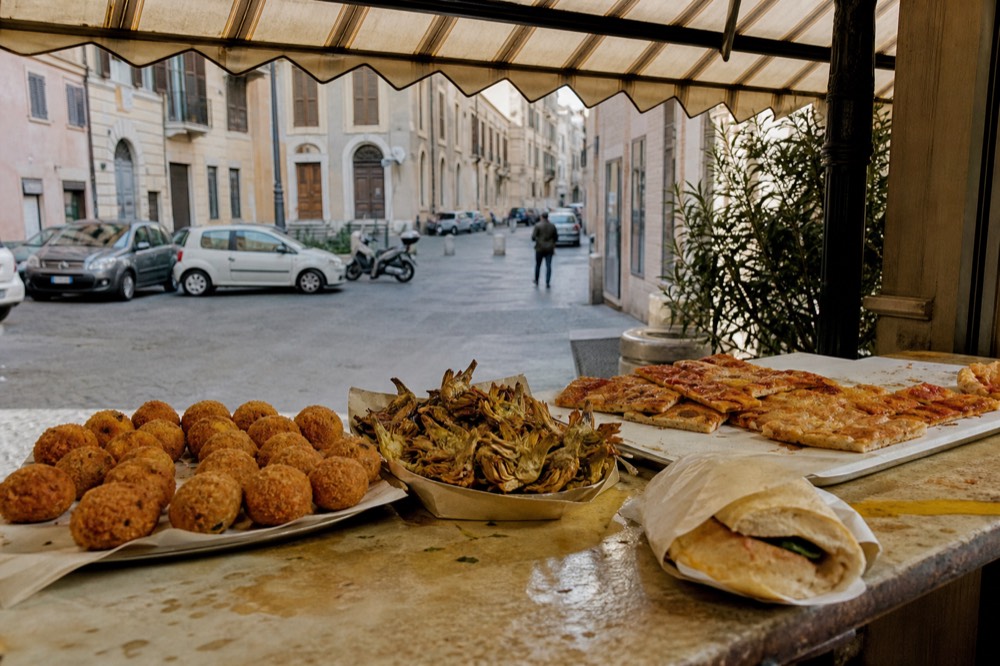 A counter of fried Roman street food, suppli and pizza slices, under a shop awning