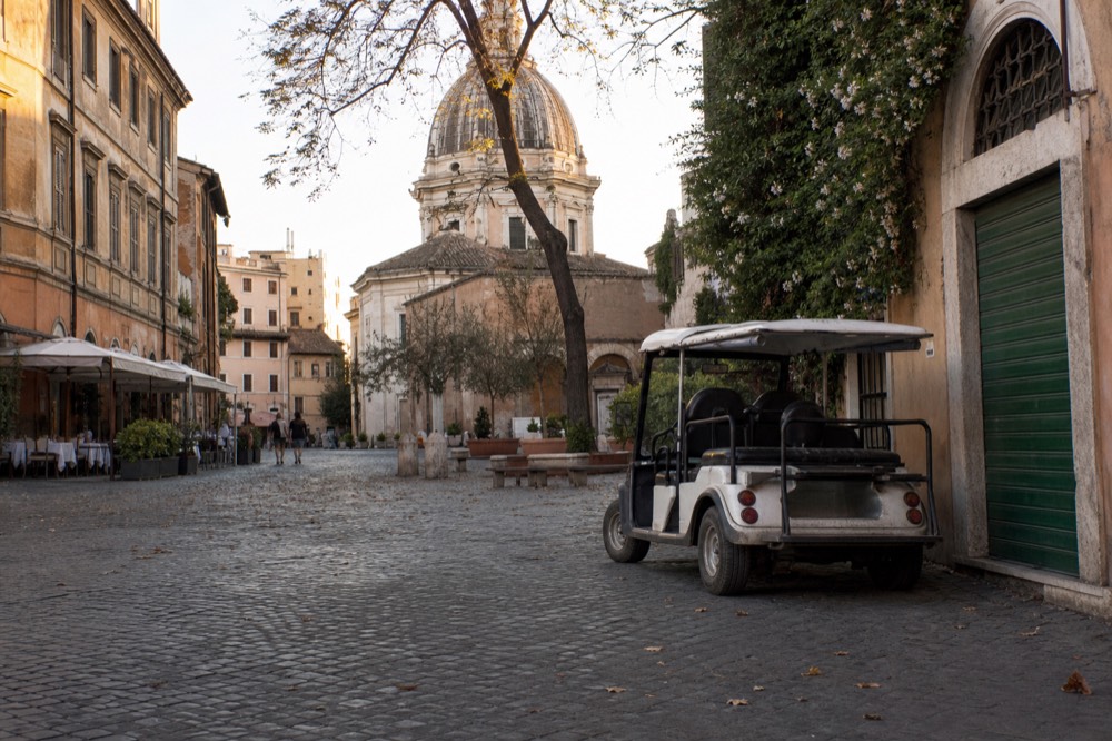 A small open golf cart parked on a cobbled Roman street near a food shop