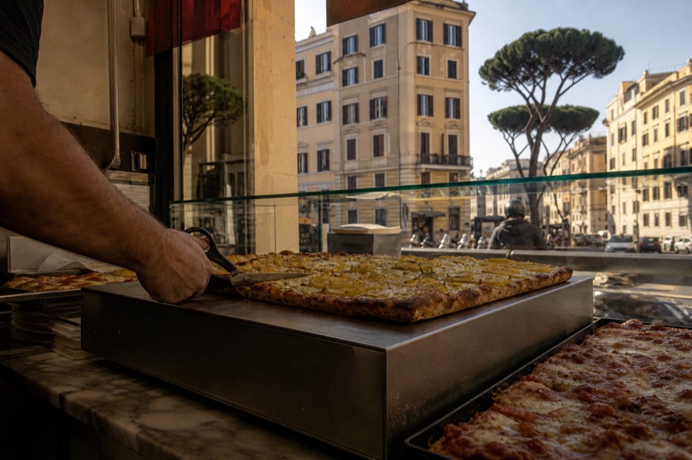 A counter worker cutting a rectangle of pizza al taglio with scissors before weighing it
