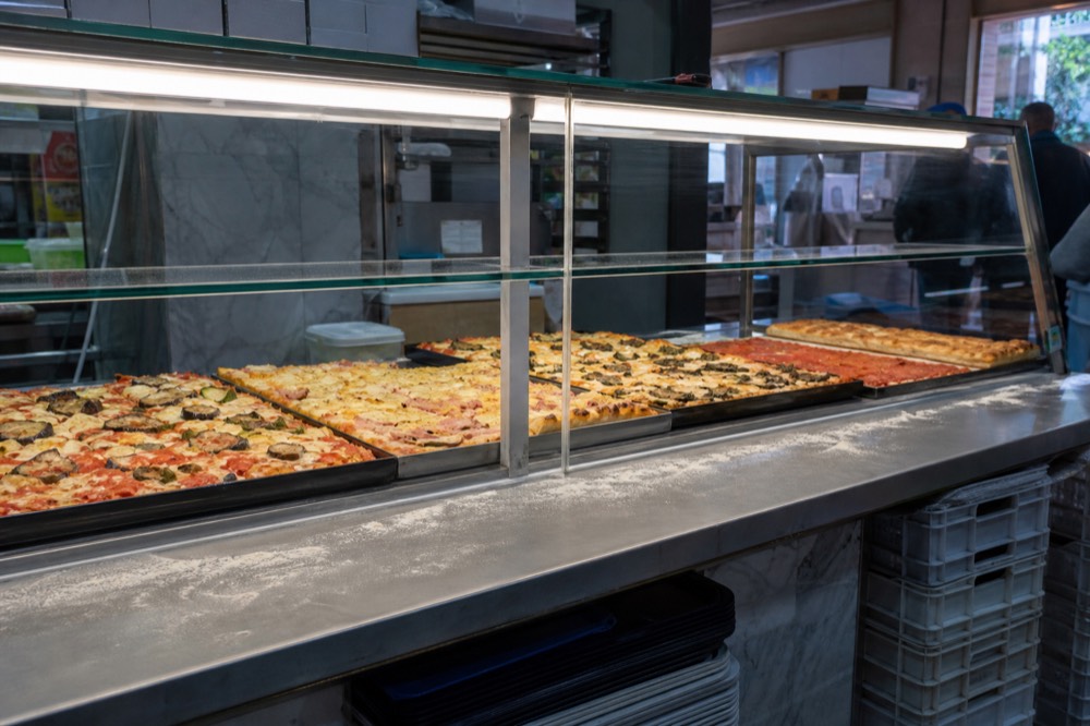 Trays of pizza al taglio under a glass counter at a bakery