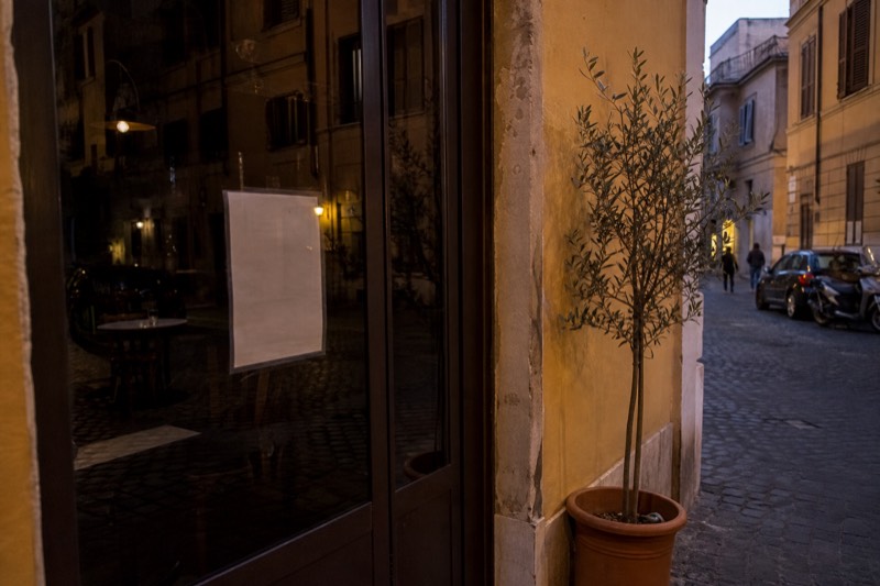 The taped glass door of a Roman trattoria showing its printed closing day, the dining room dark behind it