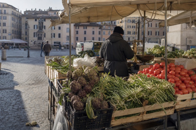 A covered market stall in Testaccio piled with artichokes, puntarelle and San Marzano tomatoes, a vendor weighing an order at the scale