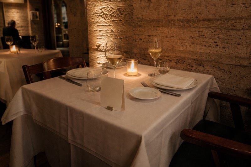 A Roman dining room at night with a table set for two, wine already poured and a small reservation card standing by the candle