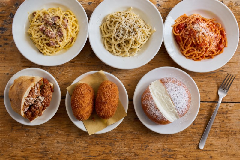 Roman classics laid out on a plain wooden counter: a plate of cacio e pepe, fried supplì in paper, a trapizzino, and a maritozzo with cream