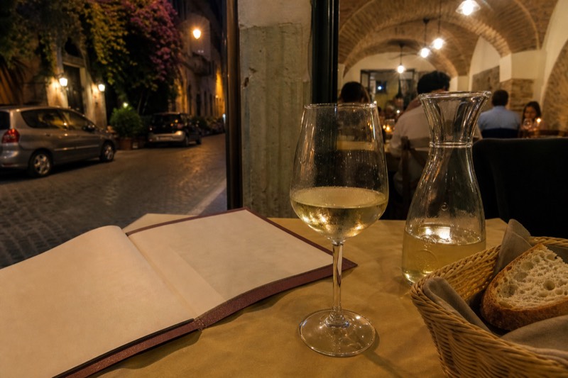 A handwritten Roman menu resting on a paper tablecloth next to a carafe of house red and a fork