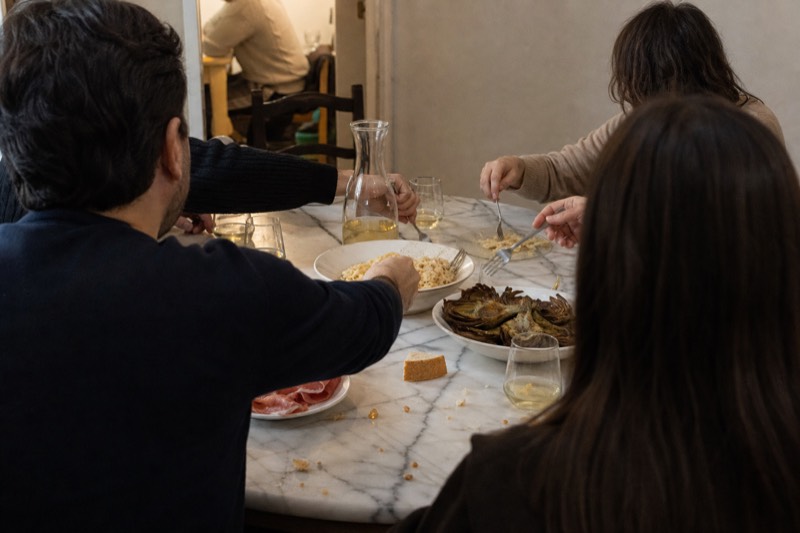 Friends at a crowded marble table in Rome reaching for bowls of cacio e pepe, a plate of fried cod and a board of cheeses, a carafe between them