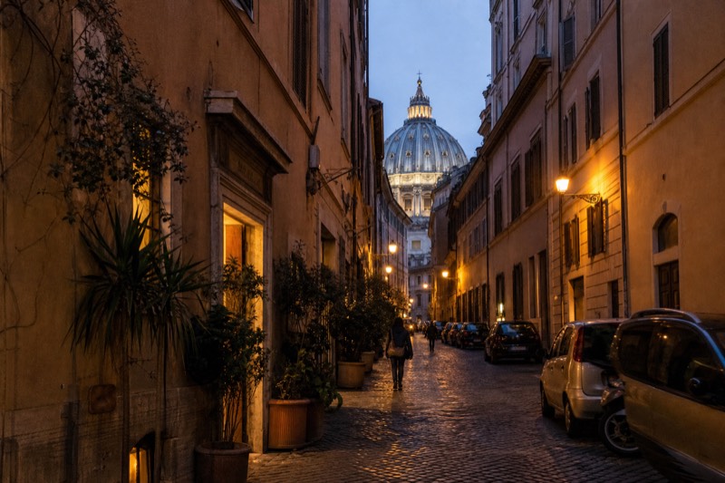 A cobbled Trastevere lane at dusk with ivy on the walls and lamps glowing in ground-floor windows, a lit dome rising beyond the rooftops
