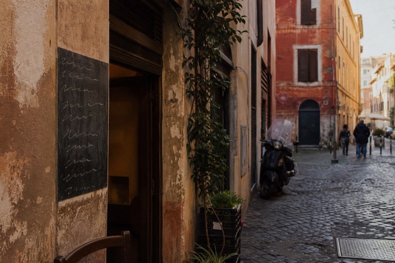 A chalkboard menu on the pavement outside a Roman trattoria listing the day's four pastas, the door half open behind it in evening light